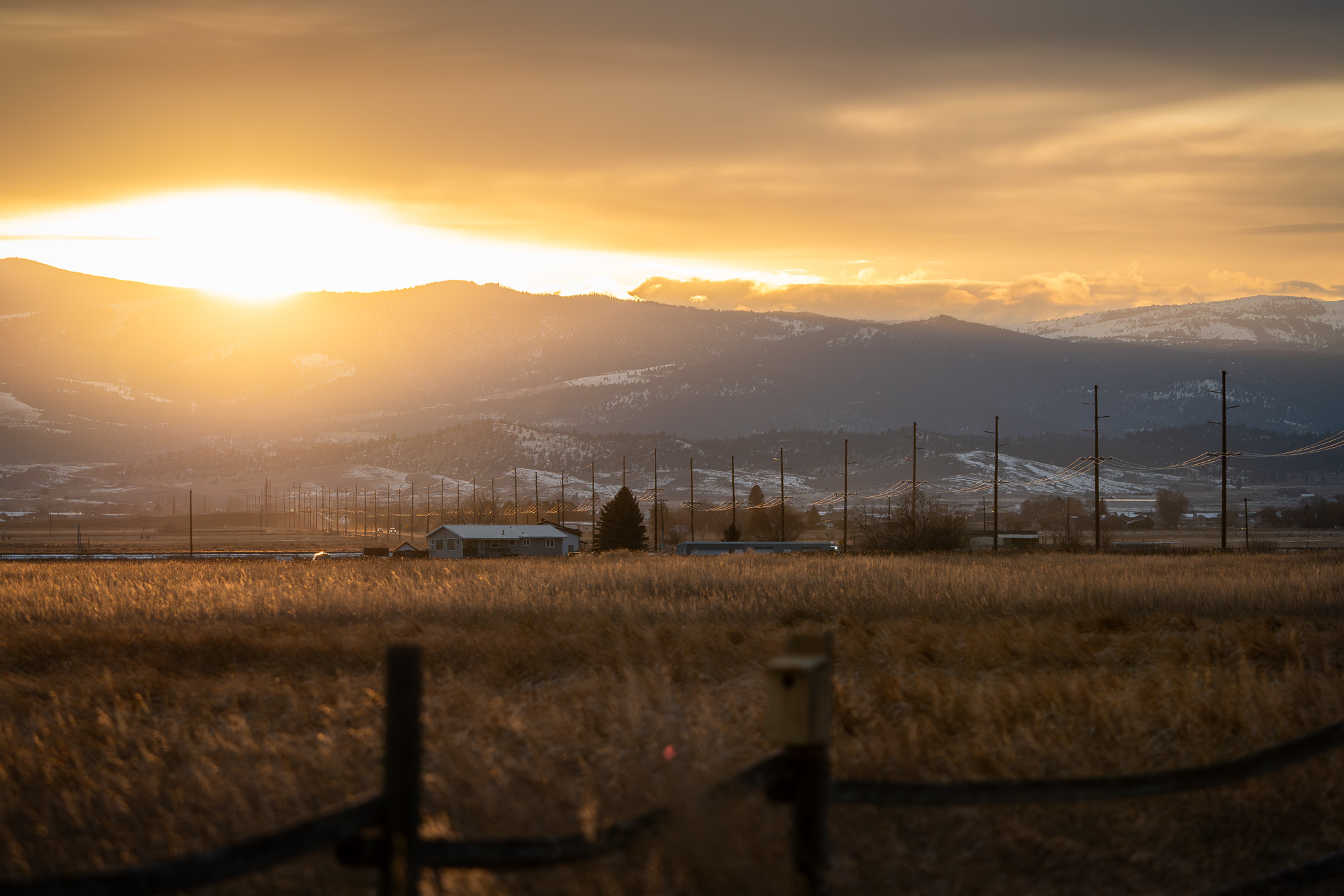 Sunset, sun is half way below a distant mountain range. a grass field is in the foreground. power poles along a road lead toward the mountains.