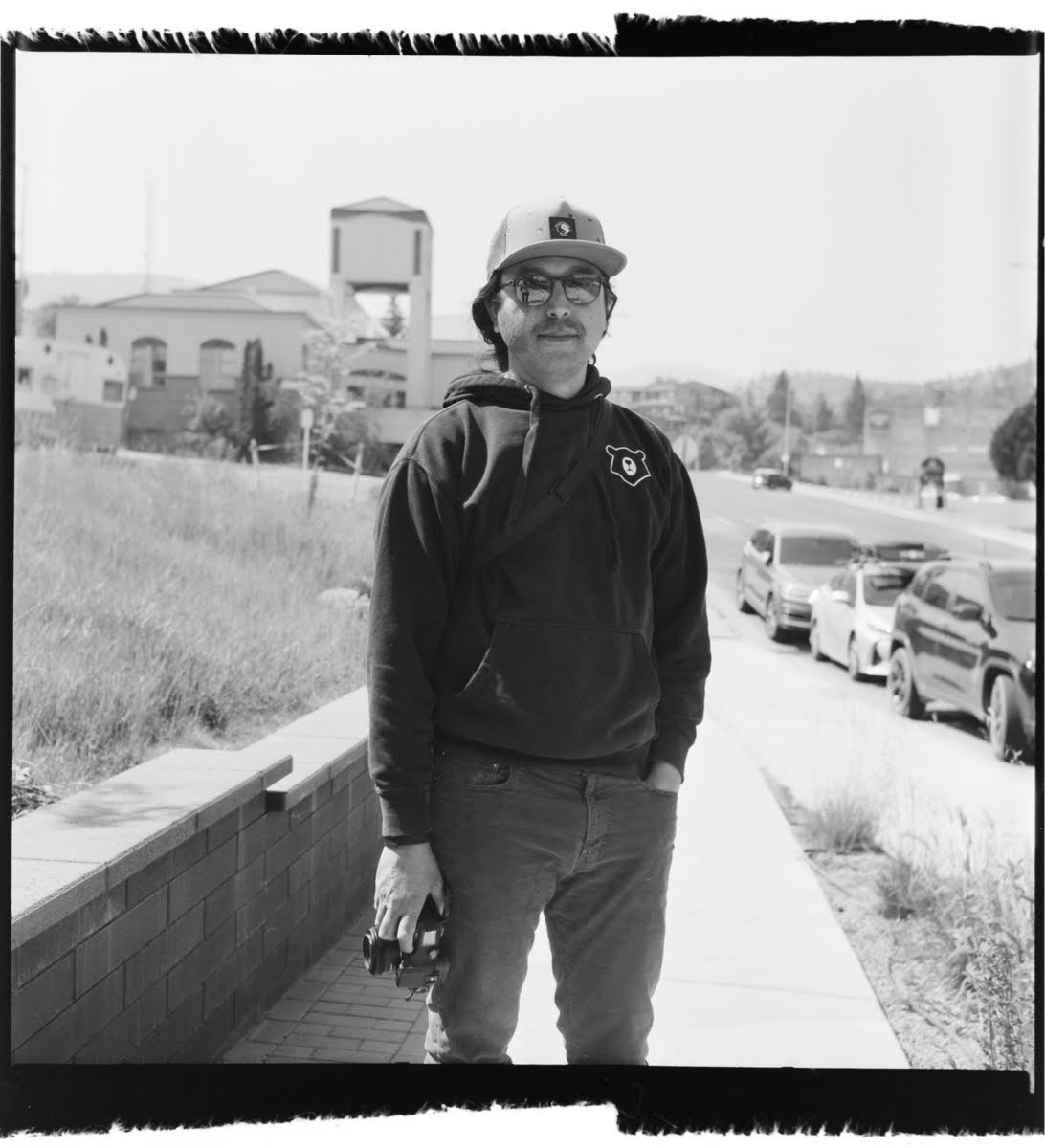 Black and white image of a man standing on a sidewalk wearing a hoodie, long pants, flat brim hat and prescription glasses. He is holding a camera in one hand at his side and looking at the camera.